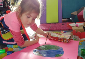 dziewczynka klęczy na kolanach i maluje pędzelkiem na kartonie umieszczonym na podłodze